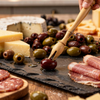 Person picking up an olive from a charcuterie board with various meats, cheeses, and olives.